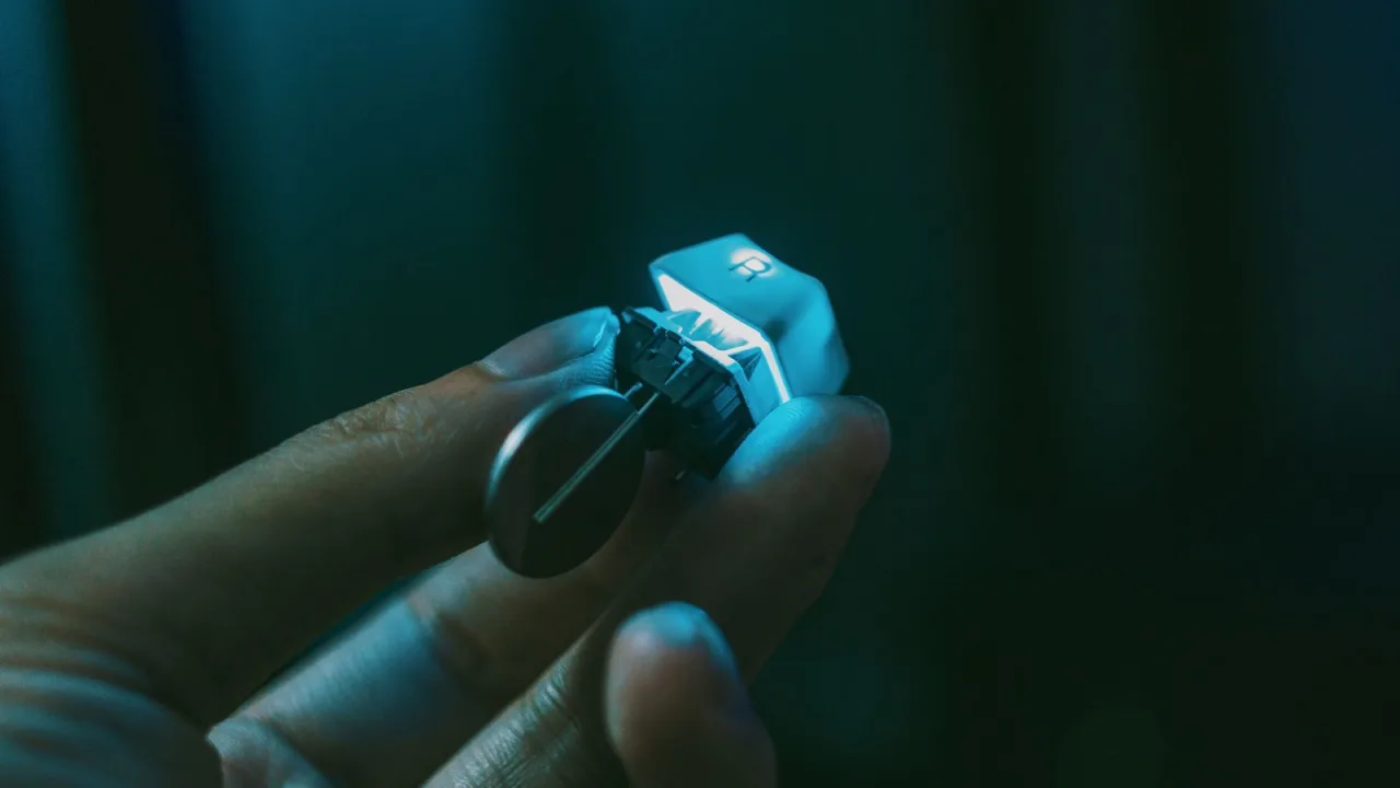 Fingers holding a single mechanical keyboard switch with a backlit keycap and a keycap puller